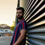 Young male model in a striped shirt posing against a modern building during sunset in Porto, Portugal.